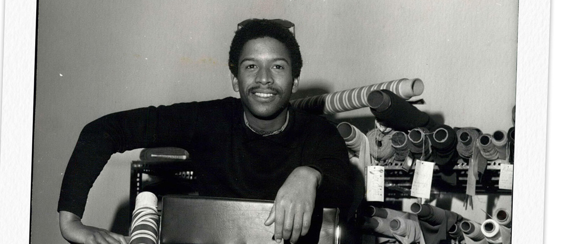 Cropped black and white image of designer Jay Jaxon sitting backward on a chair, surrounded by bolts of fabric