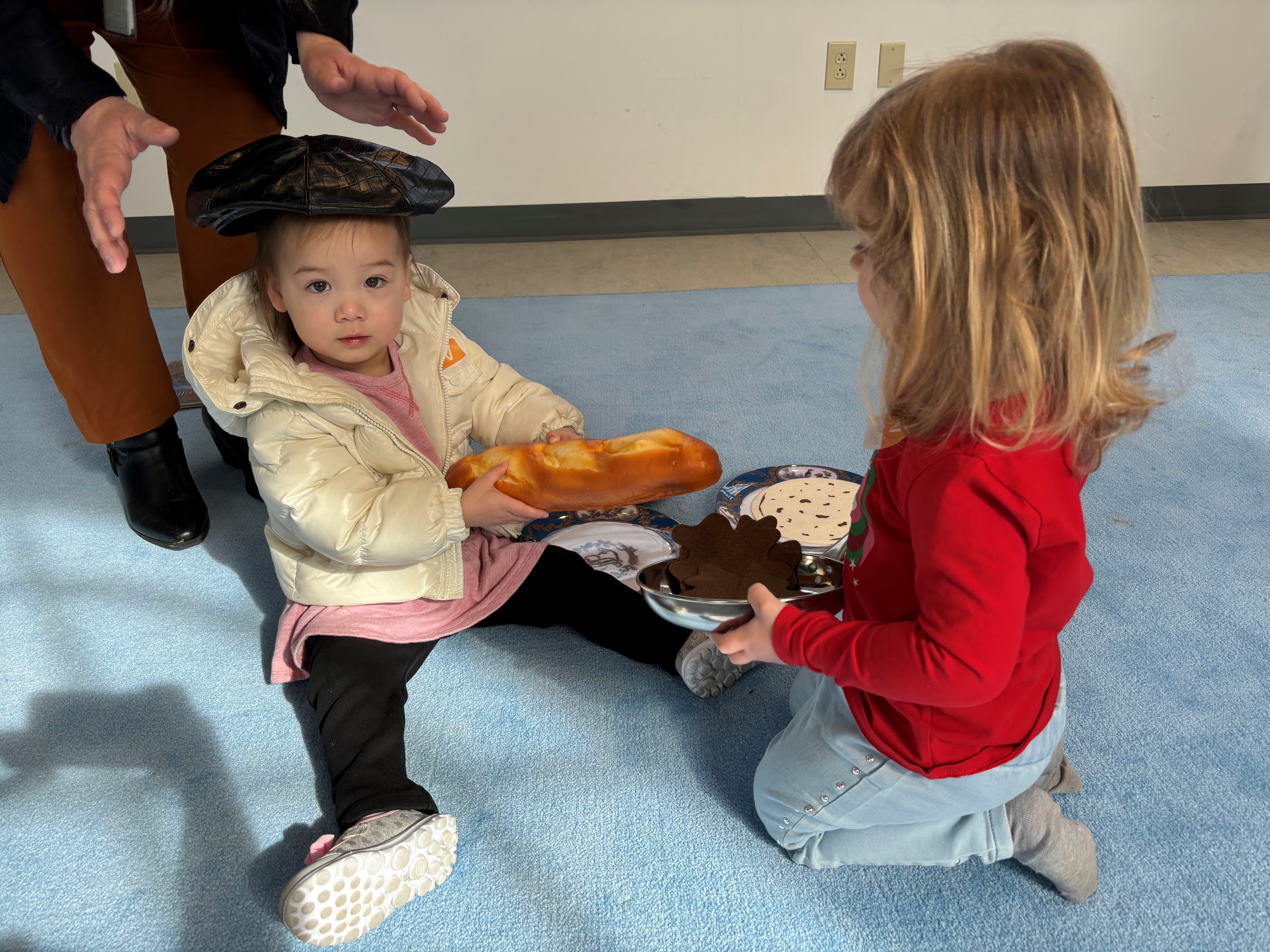 two preschoolers playing with food toys; one holds a baguette and wears a beret