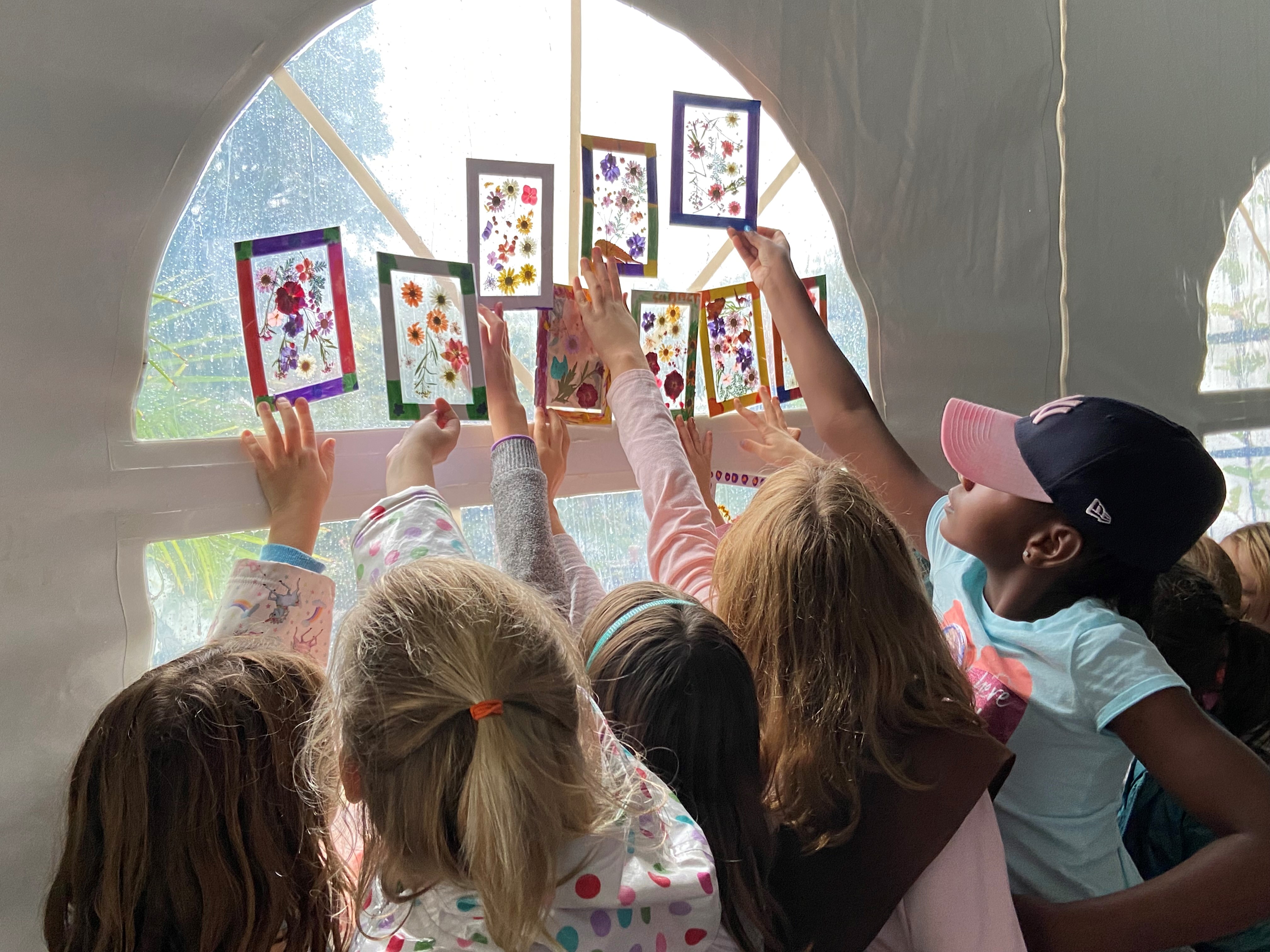 A group of Girl Scouts holding up their colorful pressed flower suncatcher art projects to catch the light streaming through a tent window.