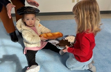 two preschoolers playing with food toys; one holds a baguette and wears a beret